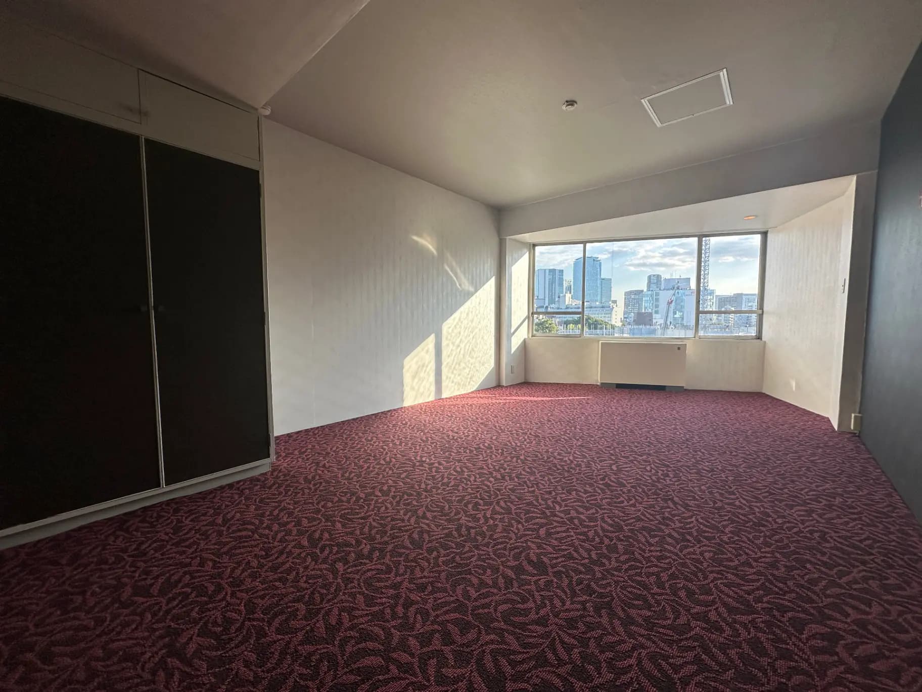 都市のスカイラインを望む広々とした部屋、赤紫色のカーペット、大きな窓、クローゼットの扉