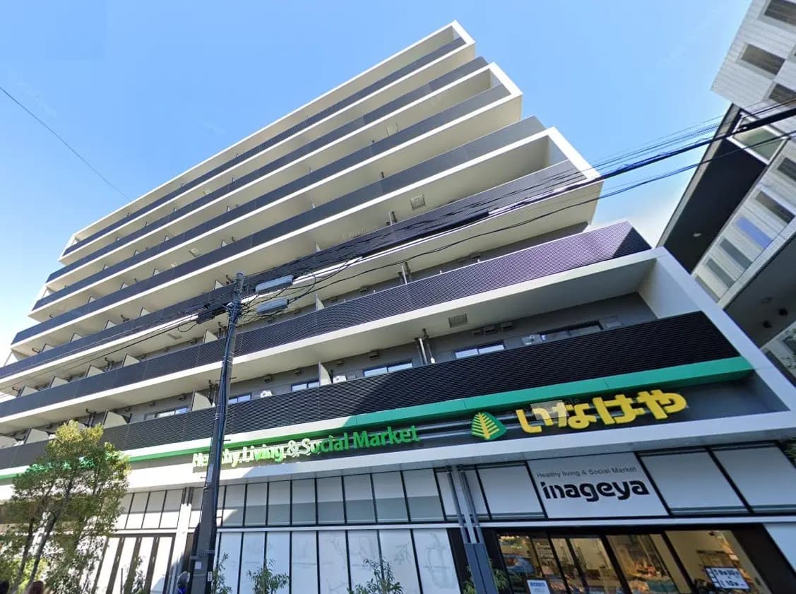 現代的な外観の多階建てアパート、1階に「いなげや」スーパーマーケットがあり、青空の下、都市の生活感が伝わる外観