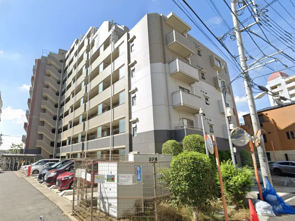 - 都会的な立地の中にある6階建てのマンション外観、青空と周囲の電線、植栽が見える景観