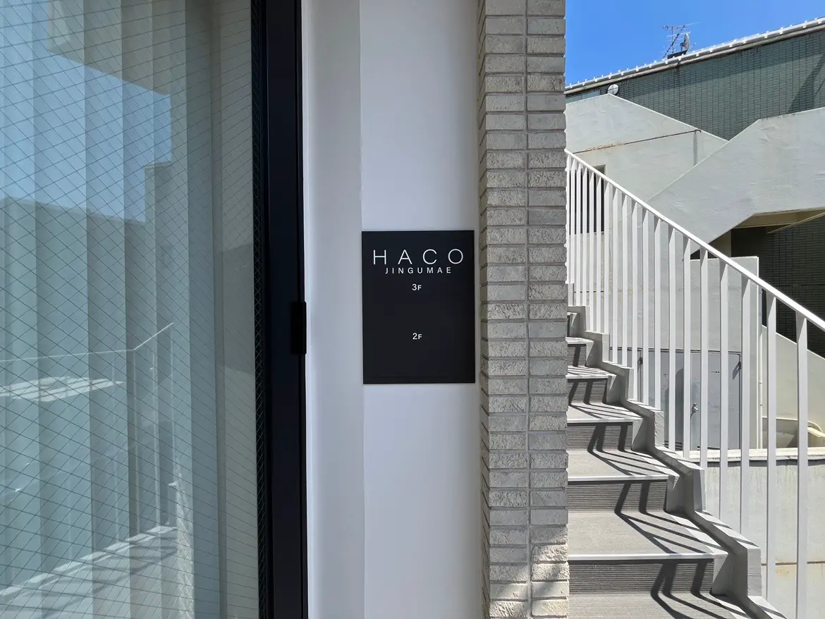- 「HACO JINGUMAE」の住所表示があるスタイリッシュなエントランス、階段と光のコントラストが印象的な建物の外観