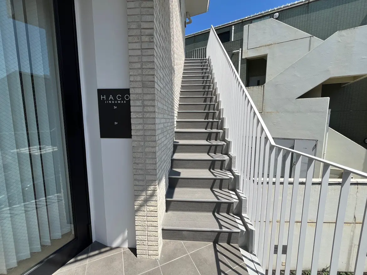 - 白い手すりがついたスロープの階段、モダンなデザインの建物外観と日差しが差し込む明るい通路の様子