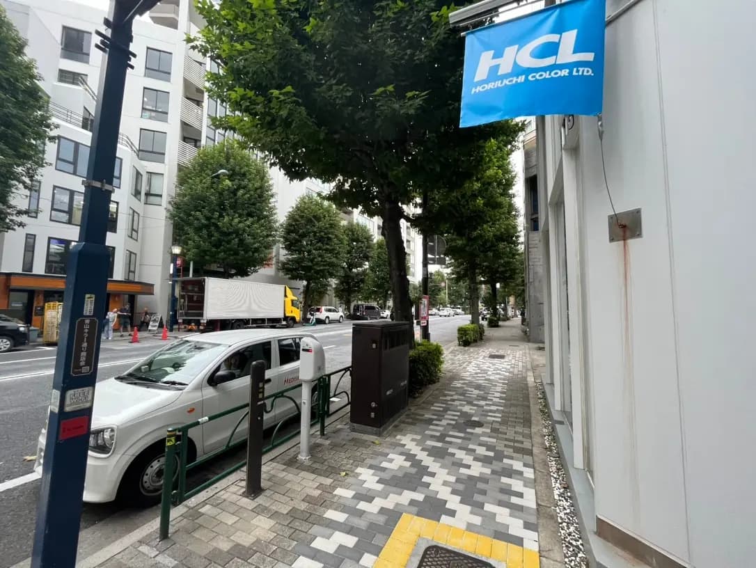 市街地の歩道と車道、障害者駐車スペースに駐車している白い車、ゲートバー、緑の木々、建物の外壁に取り付けられたHCL堀内カラーリミテッドの青い旗