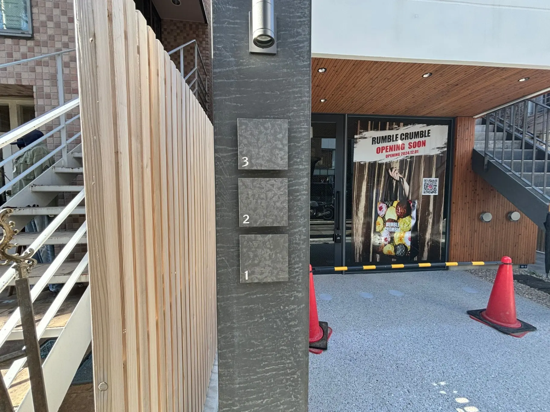 近日オープン予定の「RUMBLE CRUMBLE」と書かれた看板が掲げられた建物の入り口、階段とサンプル材木
