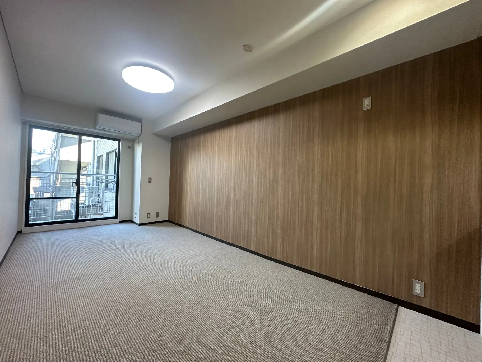 空いている賃貸アパートの部屋、明るい天井照明がついており、ベージュのカーペットと木目調の壁が特徴的なインテリア