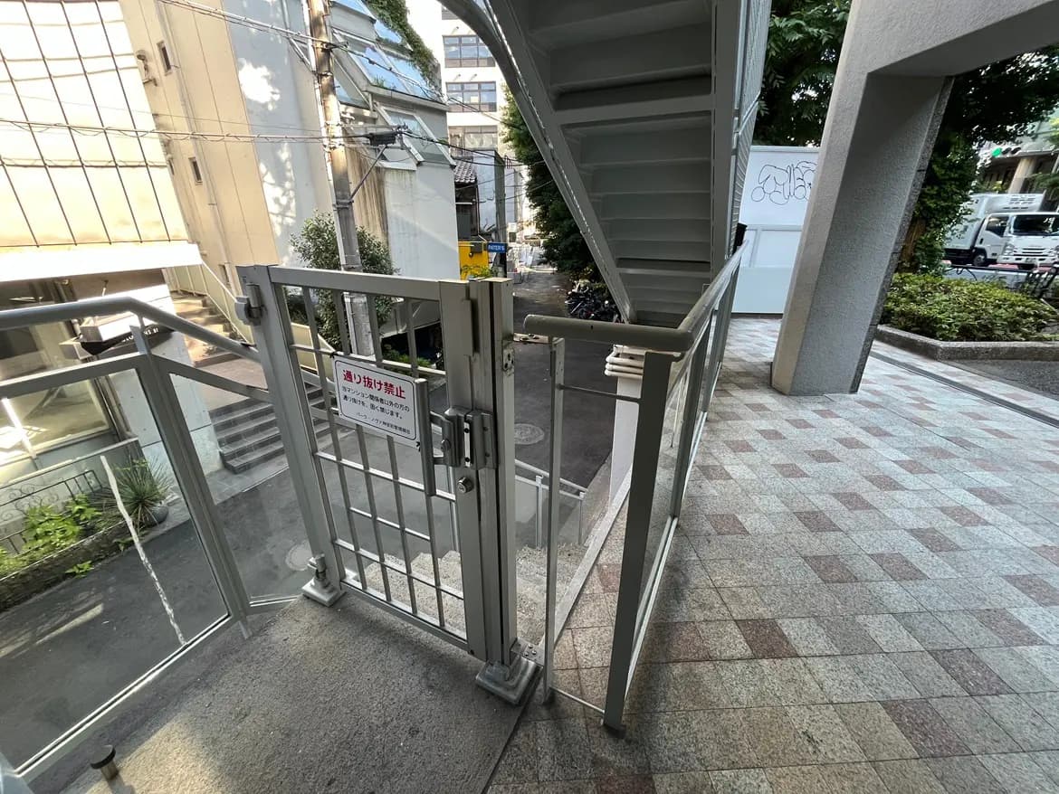 - 外階段の近くに設置された金属製のゲート、周囲は都市の景観と草木があるエントランスエリア