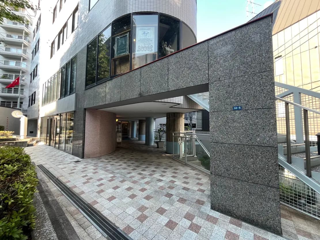 - モダンな建物のエントランス、ガラス壁とタイル舗装の道、周囲に植栽があるオープンスペース