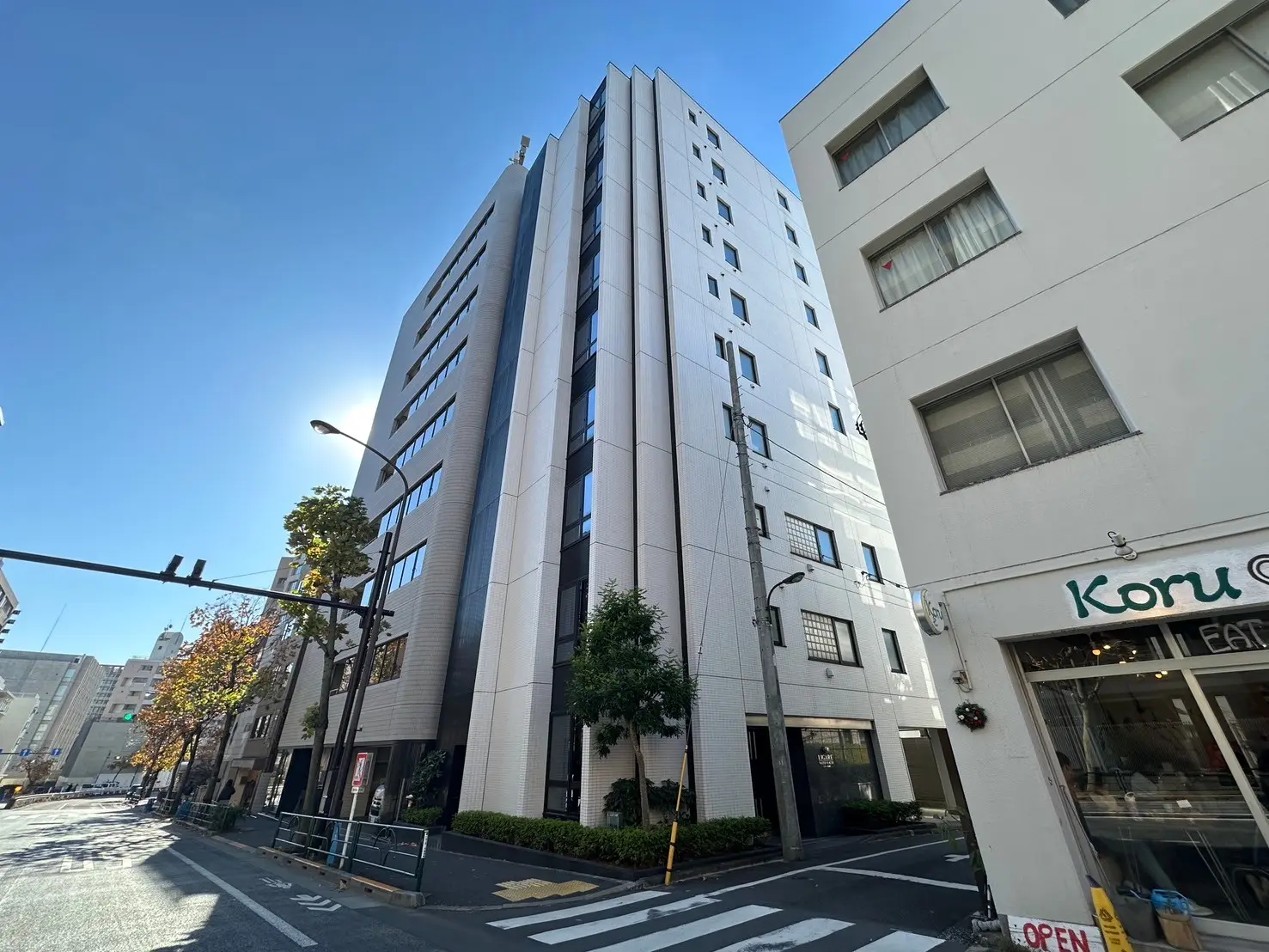 都市の通り角に建つモダンな外観のオフィスビル、晴れた日の青空と建物の影、歩道と街路樹