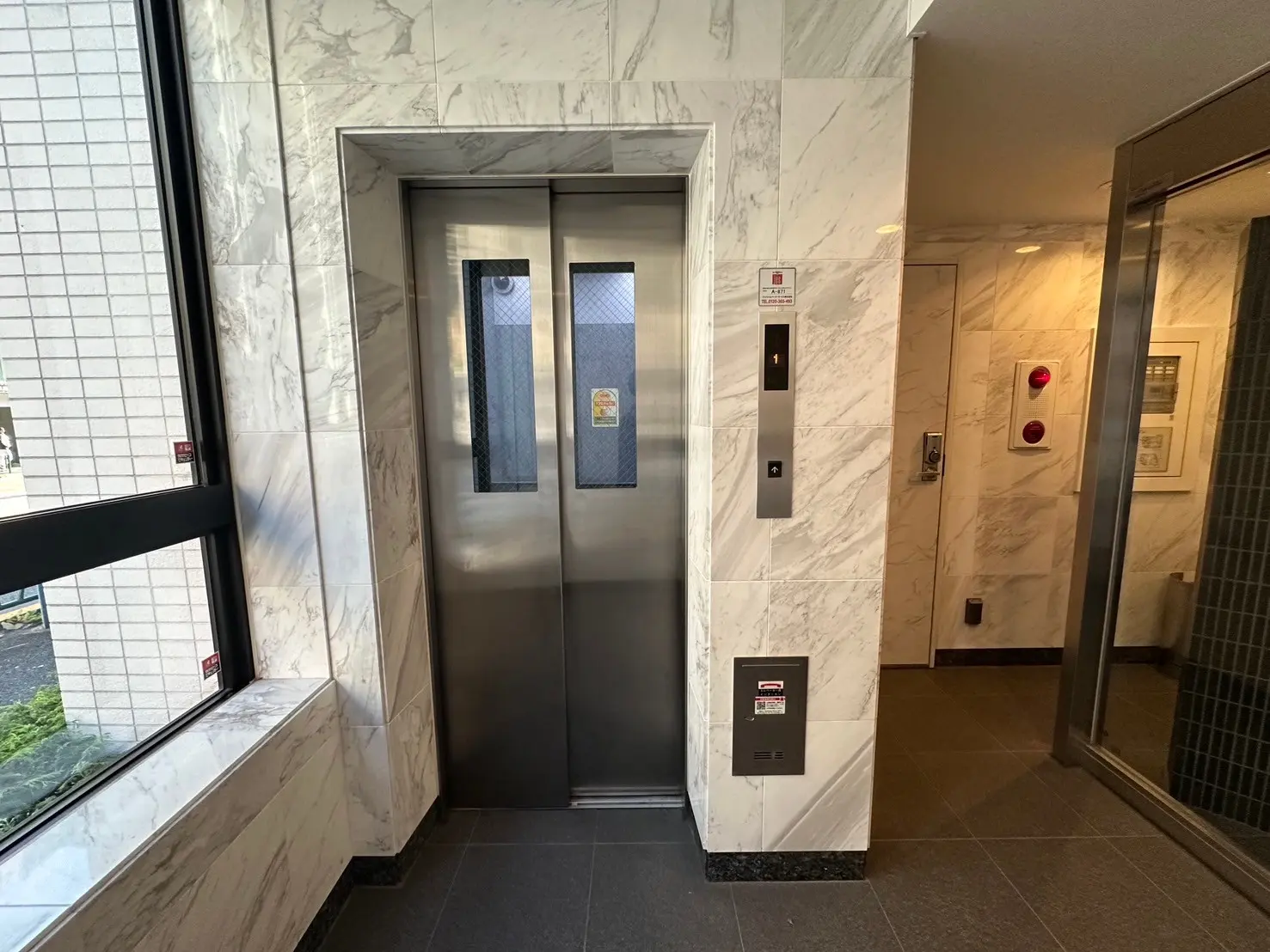 大理石調の壁面に囲まれたモダンなエレベーターホール、ガラス窓からの自然光