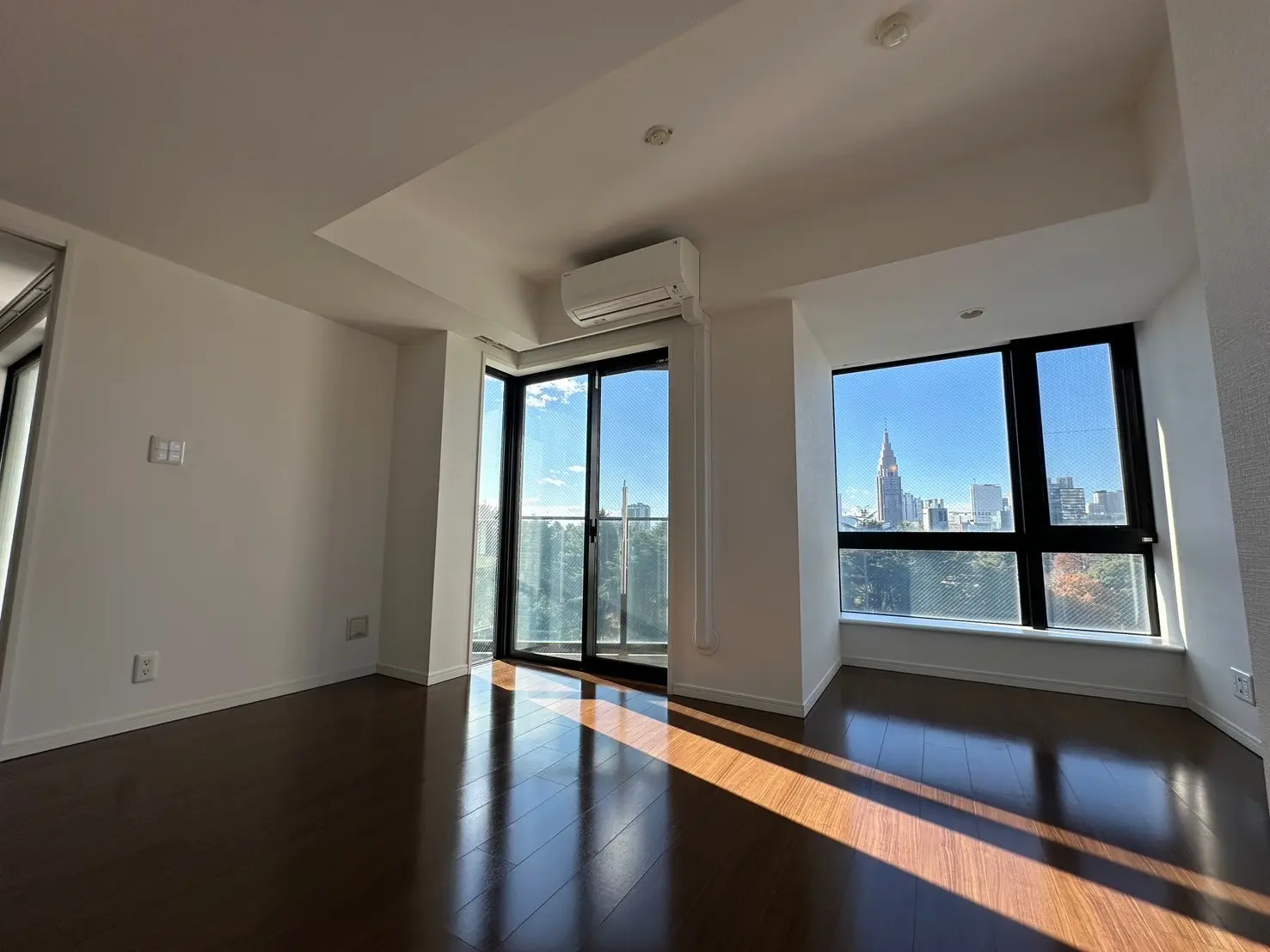 日当たりの良い空間と高層ビルの景色が見えるモダンなリビングルーム、木製フローリングと白い壁、大きな掃き出し窓