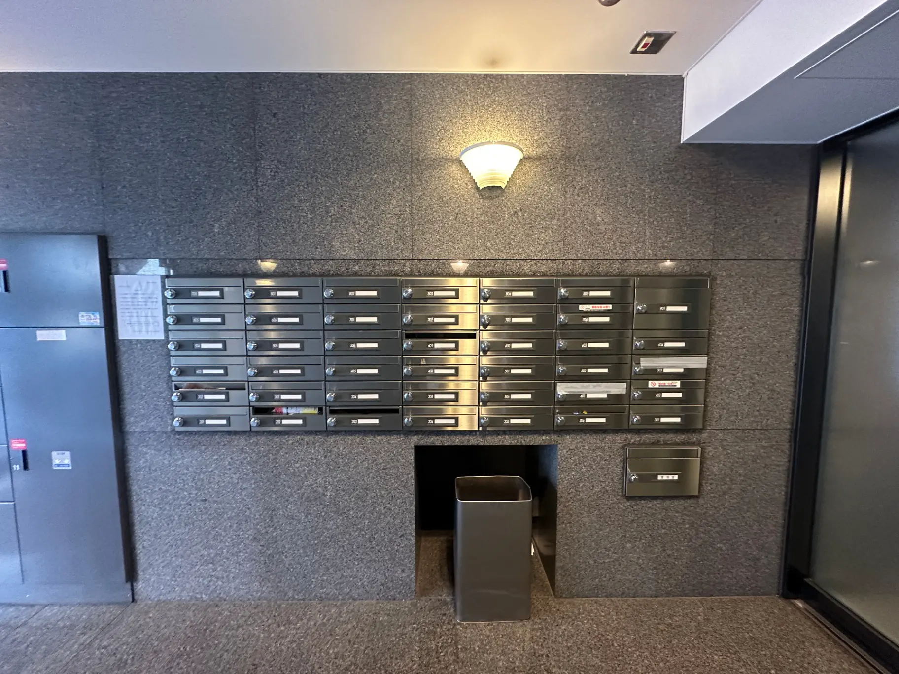 現代的なマンションのエントランスにある郵便受け、壁掛け照明、ゴミ箱、非常口案内表示