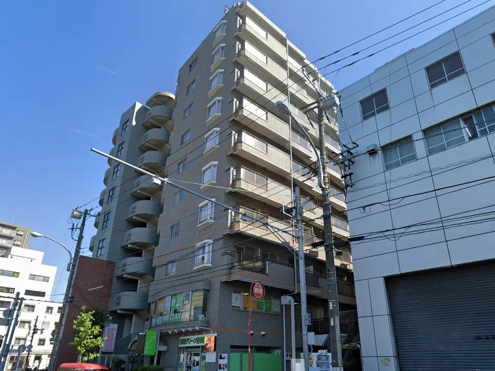 - 都市型アパートの外観、周囲には商業施設が並び、晴れた青空が広がる風景