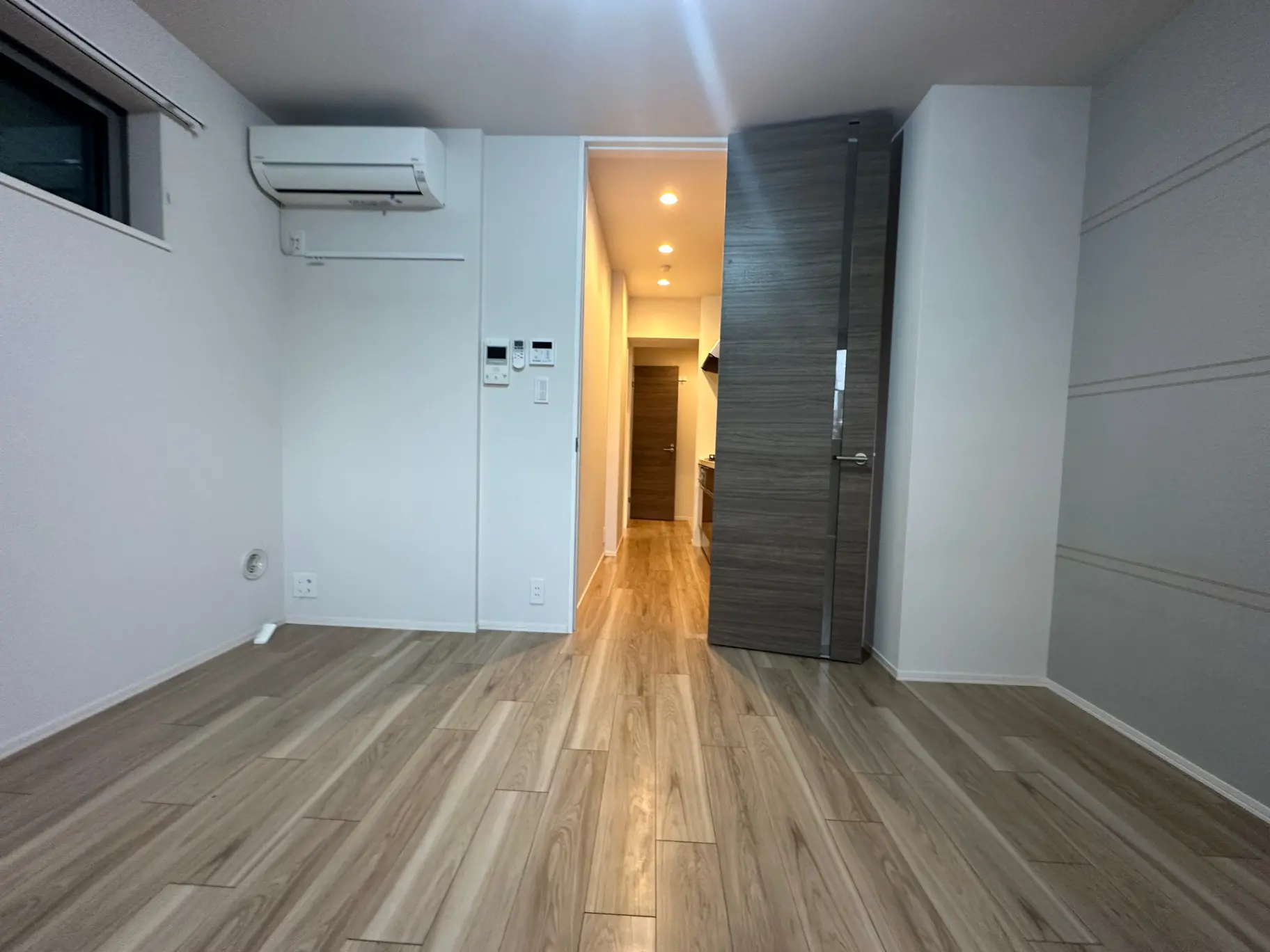 明るい木目調フローリングの空の部屋、白い壁とエアコン、奥に続く廊下が見えるインテリア