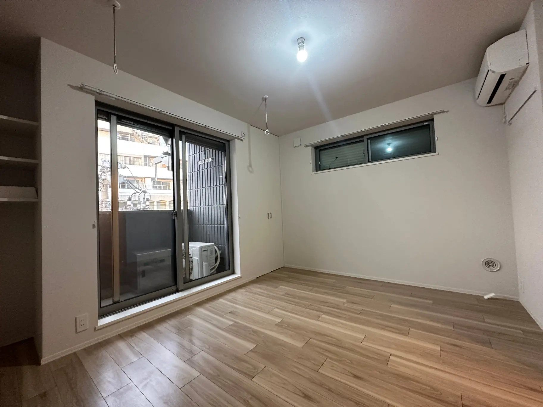 シンプルなデザインの空の部屋、木目調のフローリング、掃出し窓からの光とエアコンが設置済み