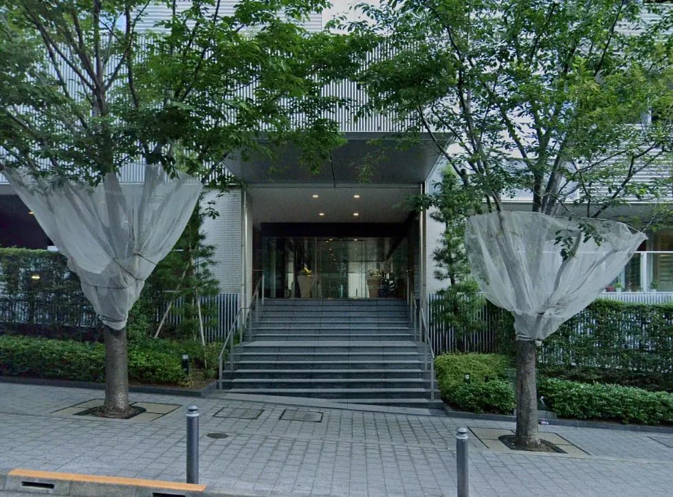 - シンプルでモダンな建物のエントランス、緑豊かな植栽と階段が特徴的で、樹木に取り付けられた風防ネットが目を引く景観