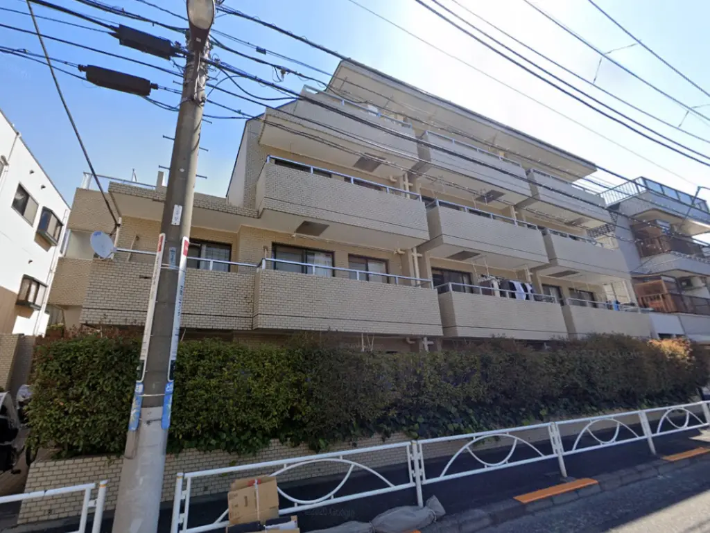 - 都市の景観に映える3階建てのマンション、バルコニーに洗濯物が干され、周囲には緑の植栽が施された外観
