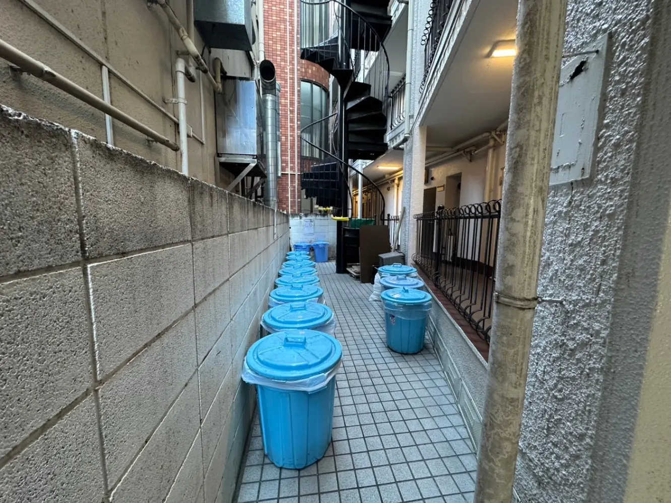 ビルの間の狭い路地、青いフタのゴミ箱が並ぶ都市風景