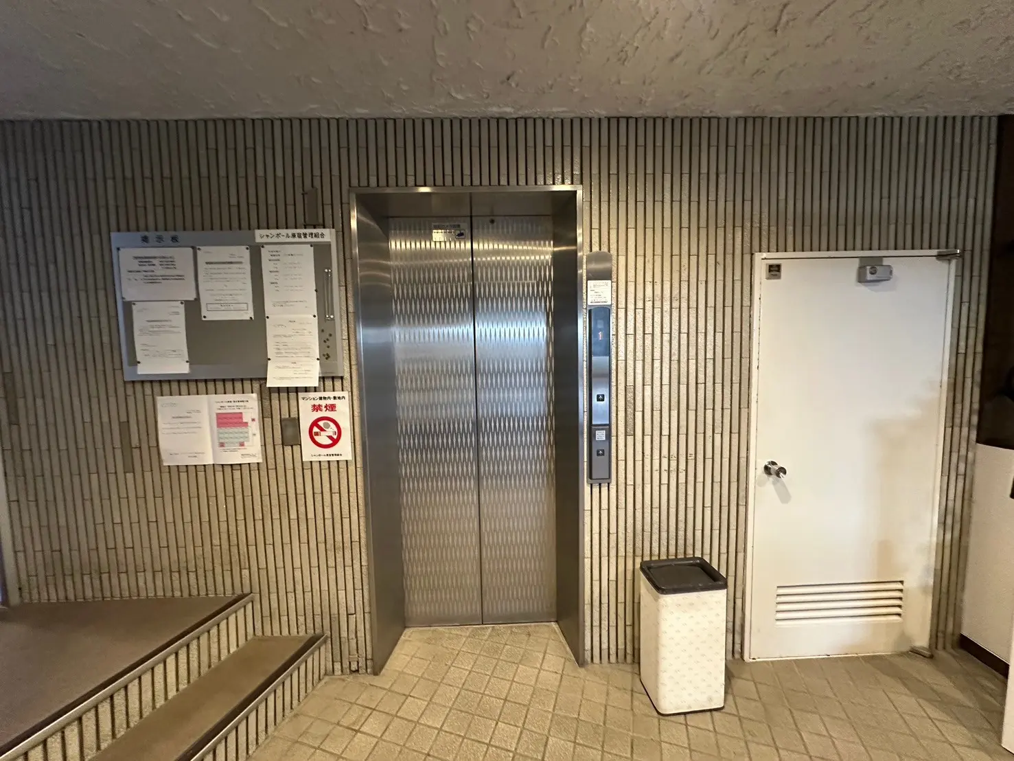 ビルのエントランスにあるステンレス製のエレベーターと掲示板、非常用の白いドアとゴミ箱があるロビー