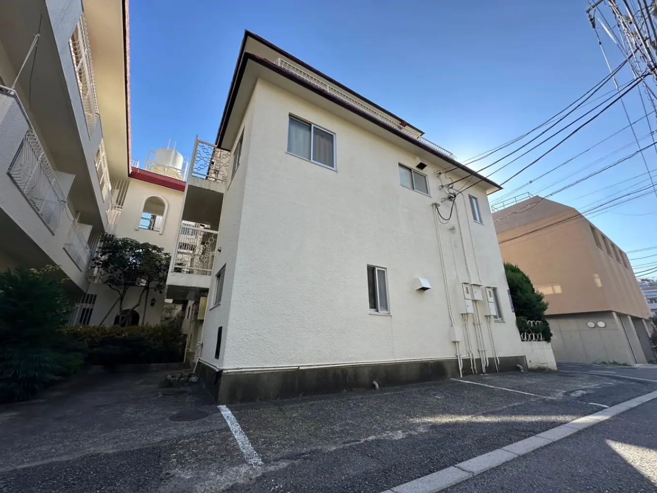 日本の住宅街にあるアパートの外観、晴れた日の青空と共に駐車場が見え、周囲には他の建物や植栽がある景色