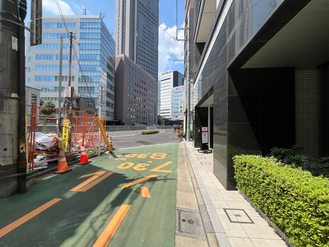 - 都市のビル街を背景にした舗装された歩道、工事現場と緑の生け垣が並ぶ風景、晴れた青空の下での街並み