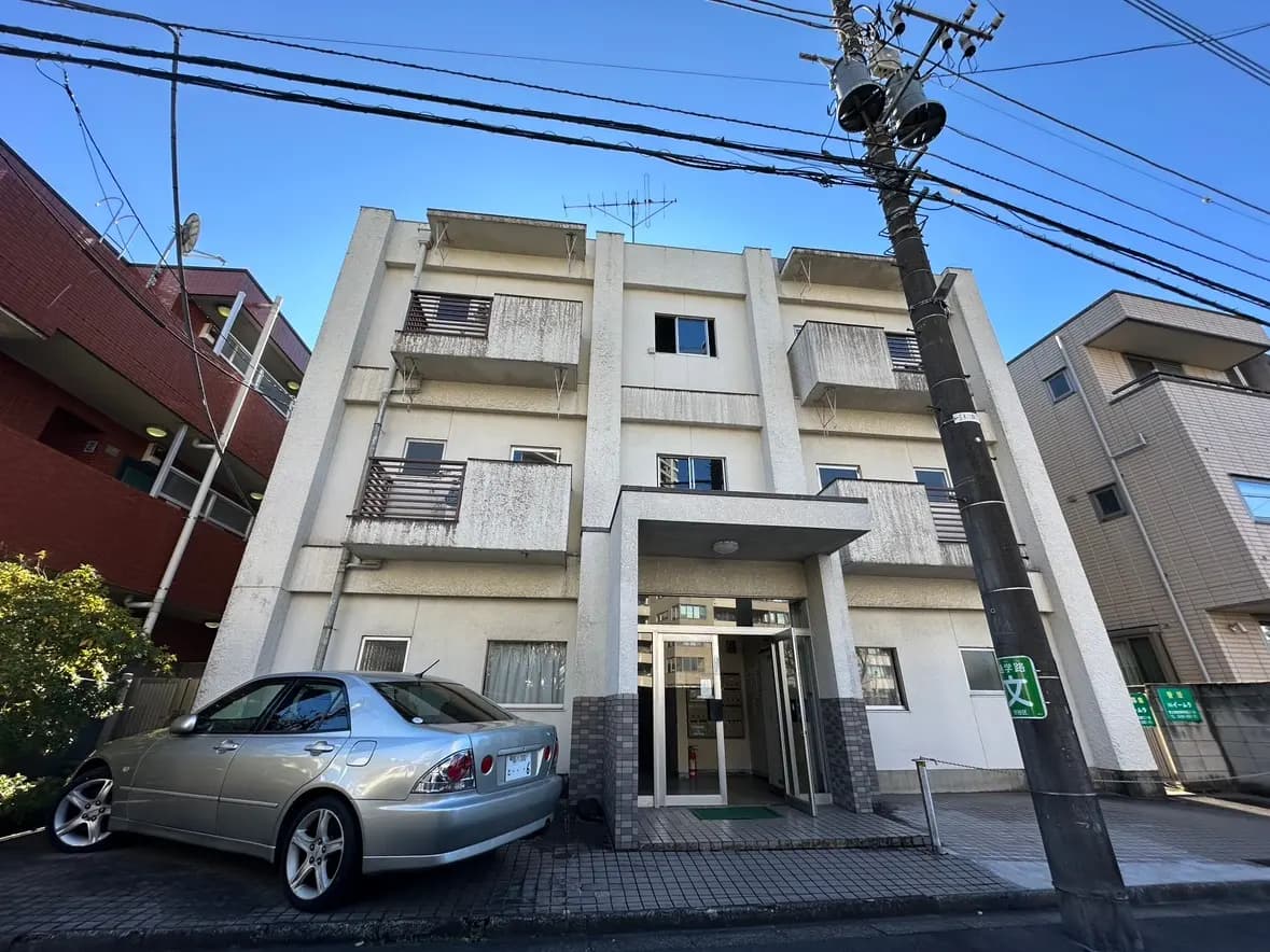 都市のアパートの外観、入口がある白い建物、前に駐車されているシルバーのセダン車