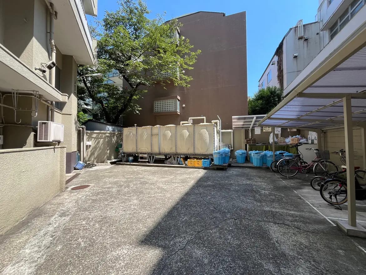 - 明るい日差しが差し込む敷地内、左側にエアコンユニット、中央に移動式トイレ、右側には青いゴミ箱と自転車が並ぶ駐輪スペースの様子
