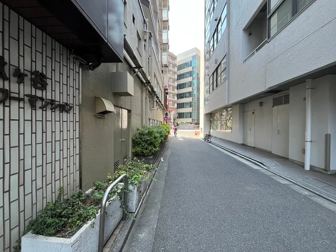 - 都会の狭い路地に面した建物、ブロック塀に取り付けられた看板と植栽、背景には高層ビルが立ち並ぶ景観