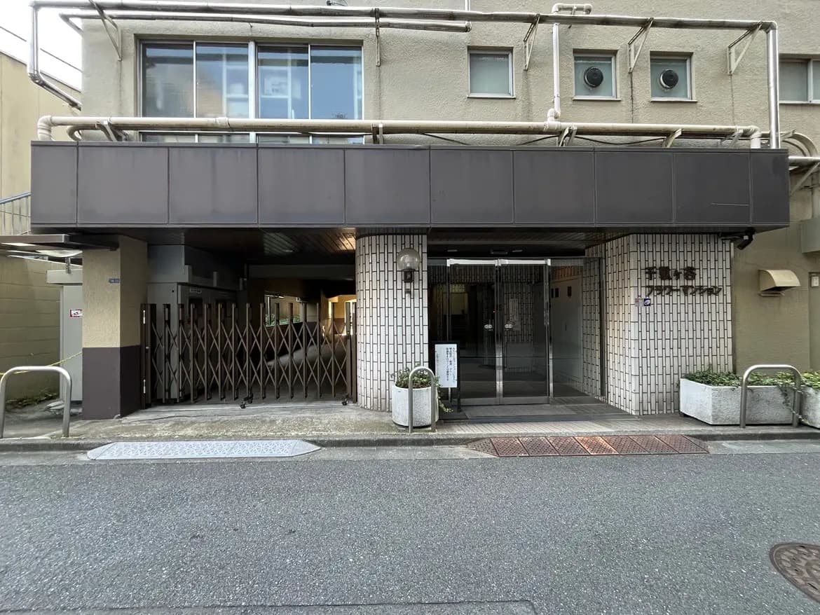 - モダンな外観のビルエントランス、特徴的なスチールの屋根とガラスドア、周囲は舗装された道路と植栽で囲まれた施設の入り口