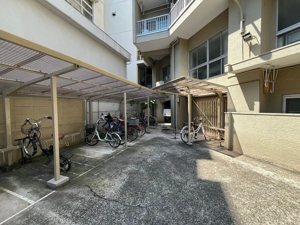 - 自転車が並ぶ屋根付きの駐輪場、周囲に高い建物がある日差しの中での風景