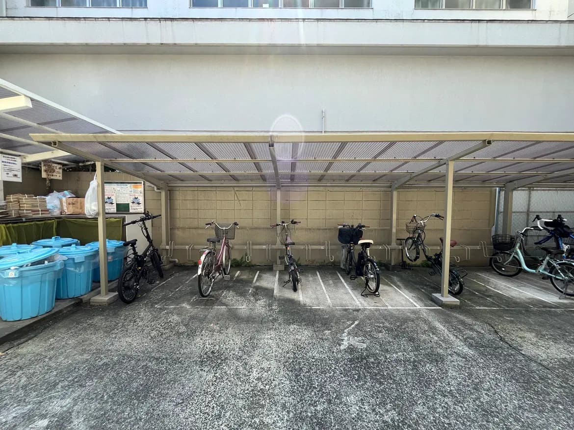 - 駐輪場の様子、自転車が並び、青いゴミ箱が置かれた屋根付きのスペース