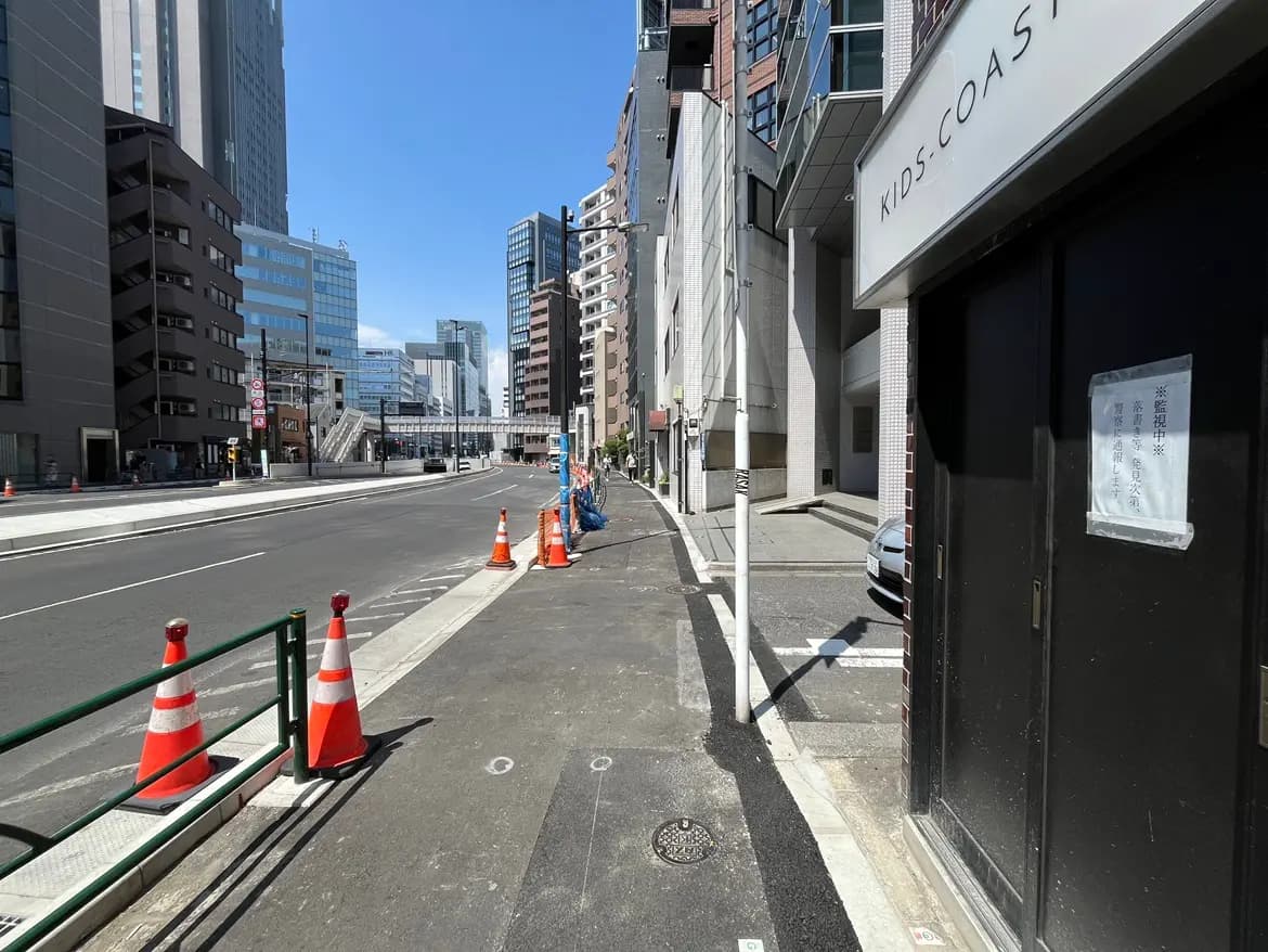 - 都市の中心にある通りの風景、青空の下に立つ高層ビルと工事中の施設、交通規制のためのコーンが並ぶ歩道。