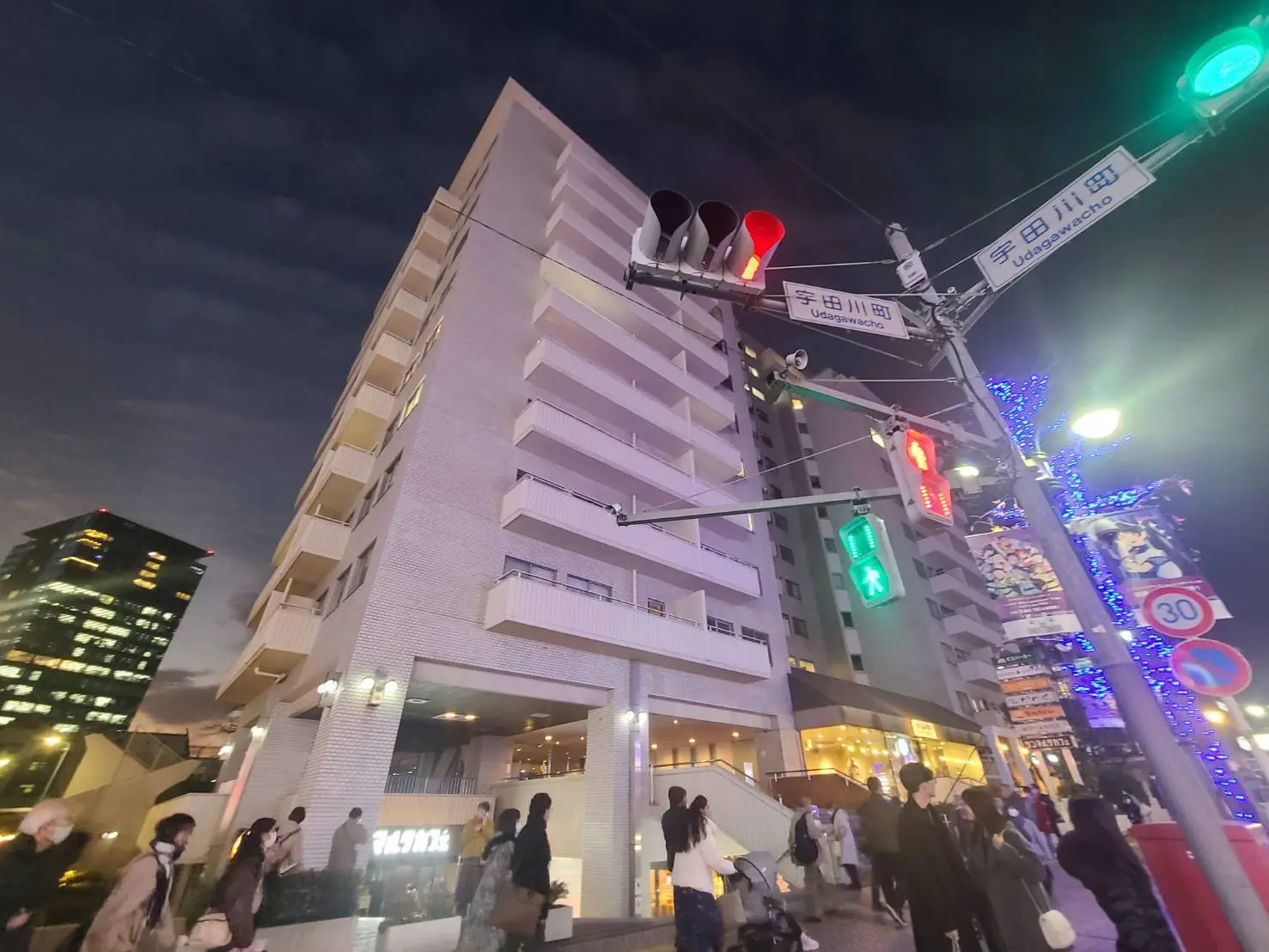 夕暮れ時の都市風景、多くの人々が歩道を歩いている、交差点にある信号機と街灯、背景にはビルのライトアップ