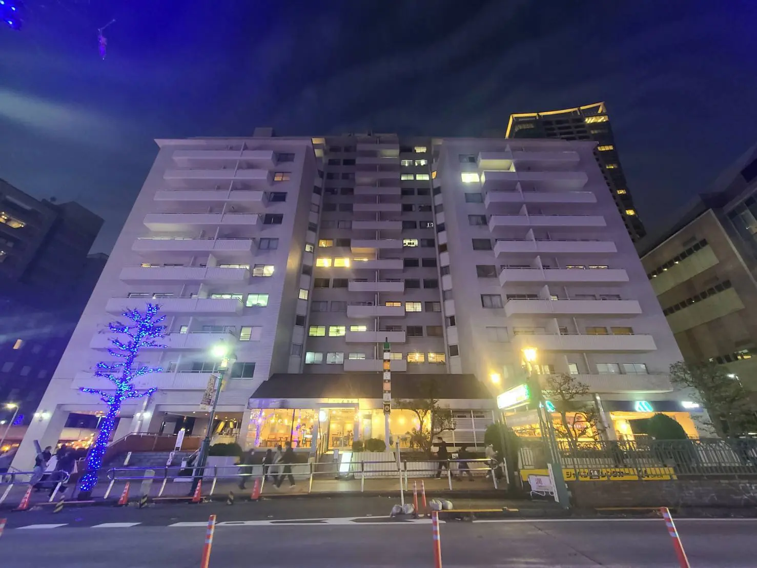 夜景が映える都市のマンションビル、イルミネーションで飾られた木、周辺の建物と通り抜ける人々