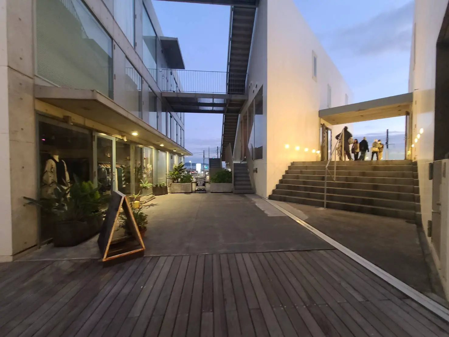 夕暮れ時のモダンな商業施設の屋外通路、照明が灯る階段とショップ、人々が集うエントランスエリア
