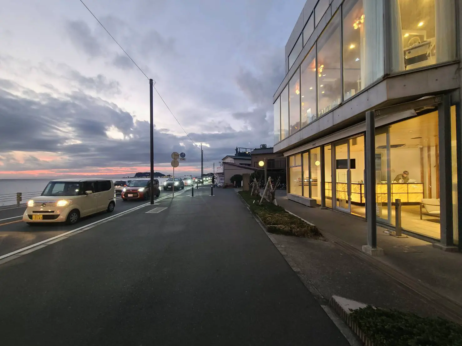 夕暮れ時の海岸通りとモダンなカフェの外観、温かい光が窓から漏れる風景