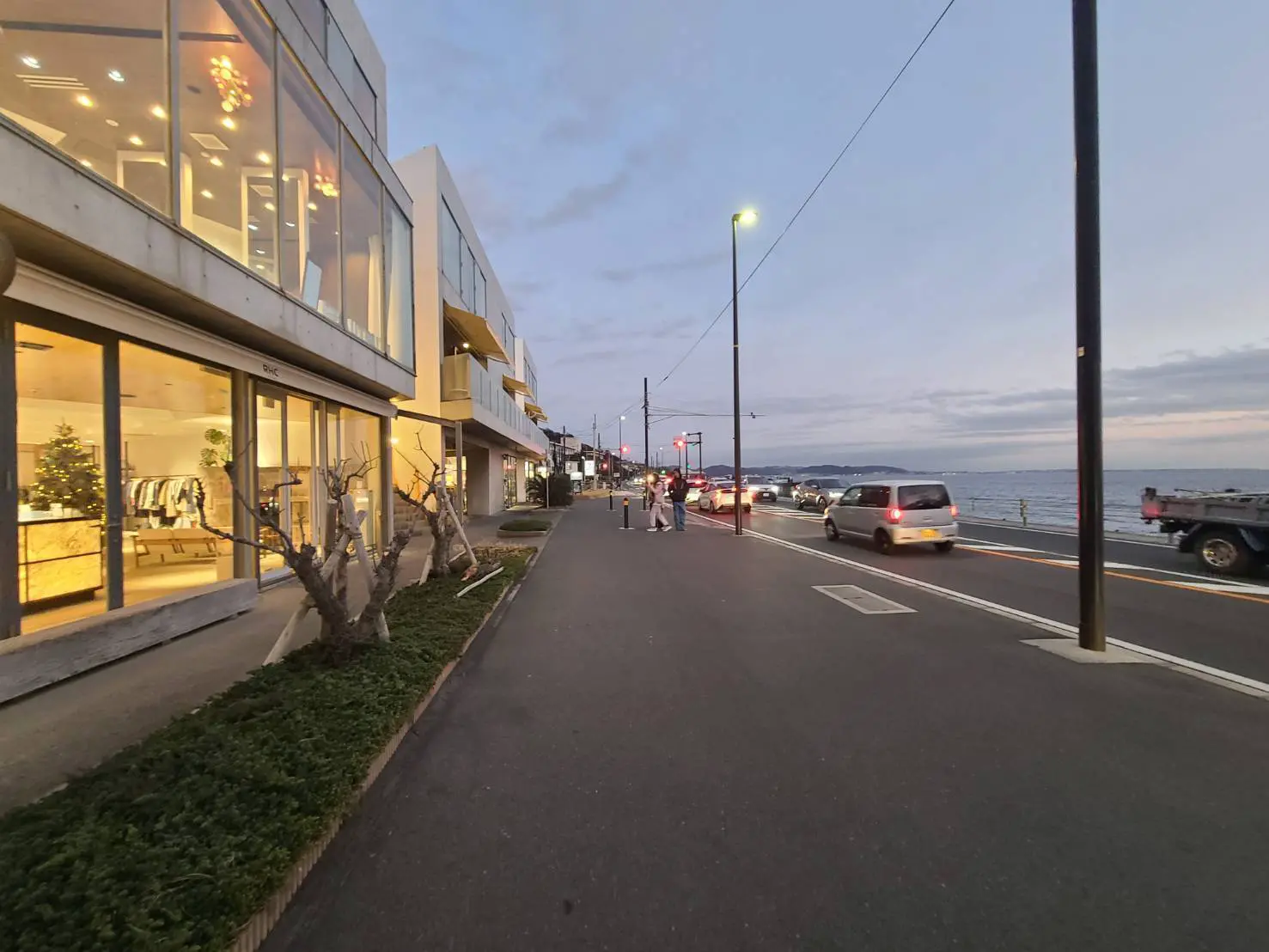 海沿いの遊歩道と商店街、夕暮れ時の風景、人々が散歩する様子とライトアップされたショーウィンドウ