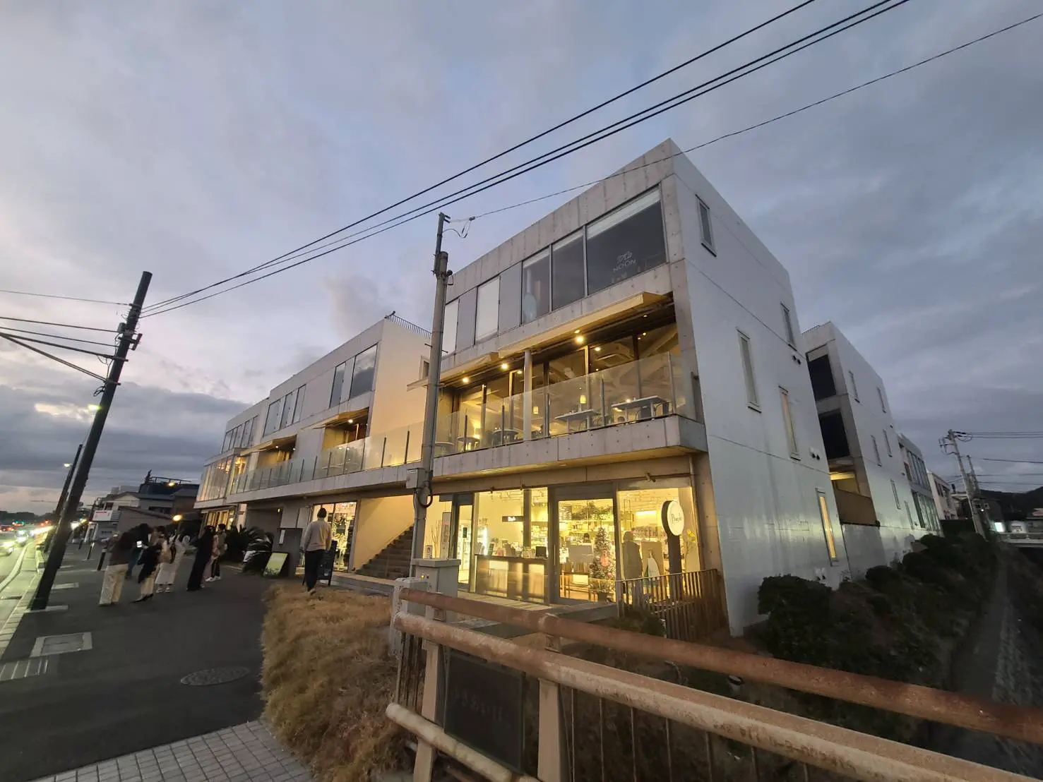夕暮れ時のモダンなビルディング、一階には照明が灯る店舗、人々が歩道を歩くシティライフ