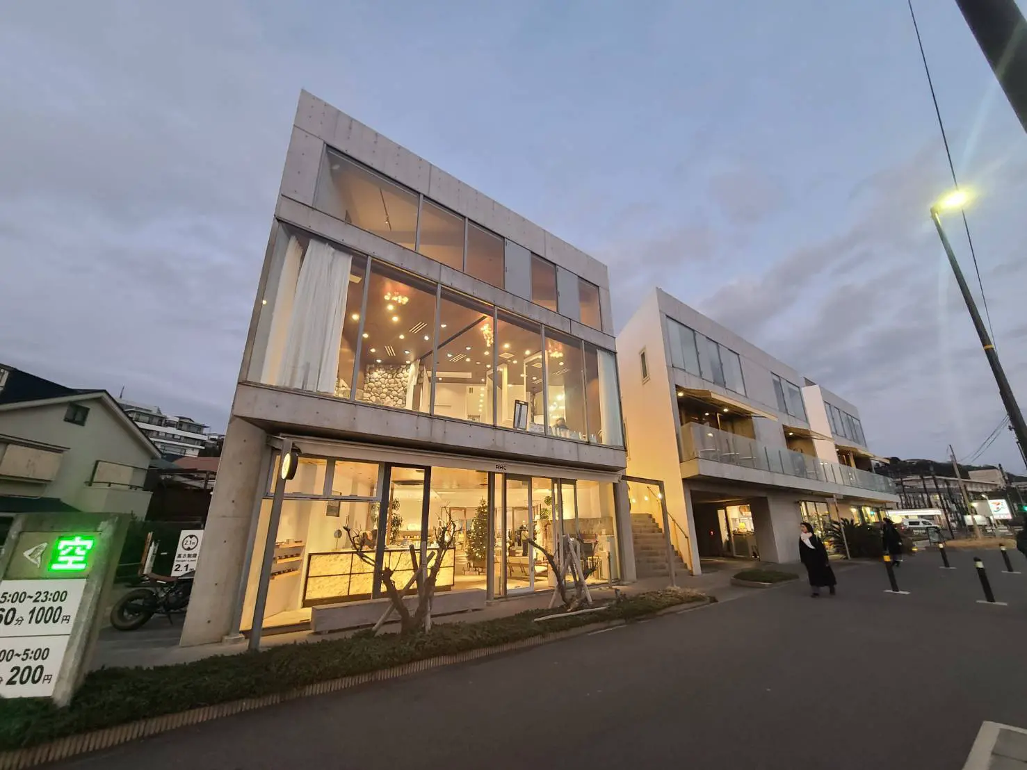 夕暮れ時に撮影されたモダンな外観の複数階建てビル、ガラス張りで照明が灯るインテリアが見える
