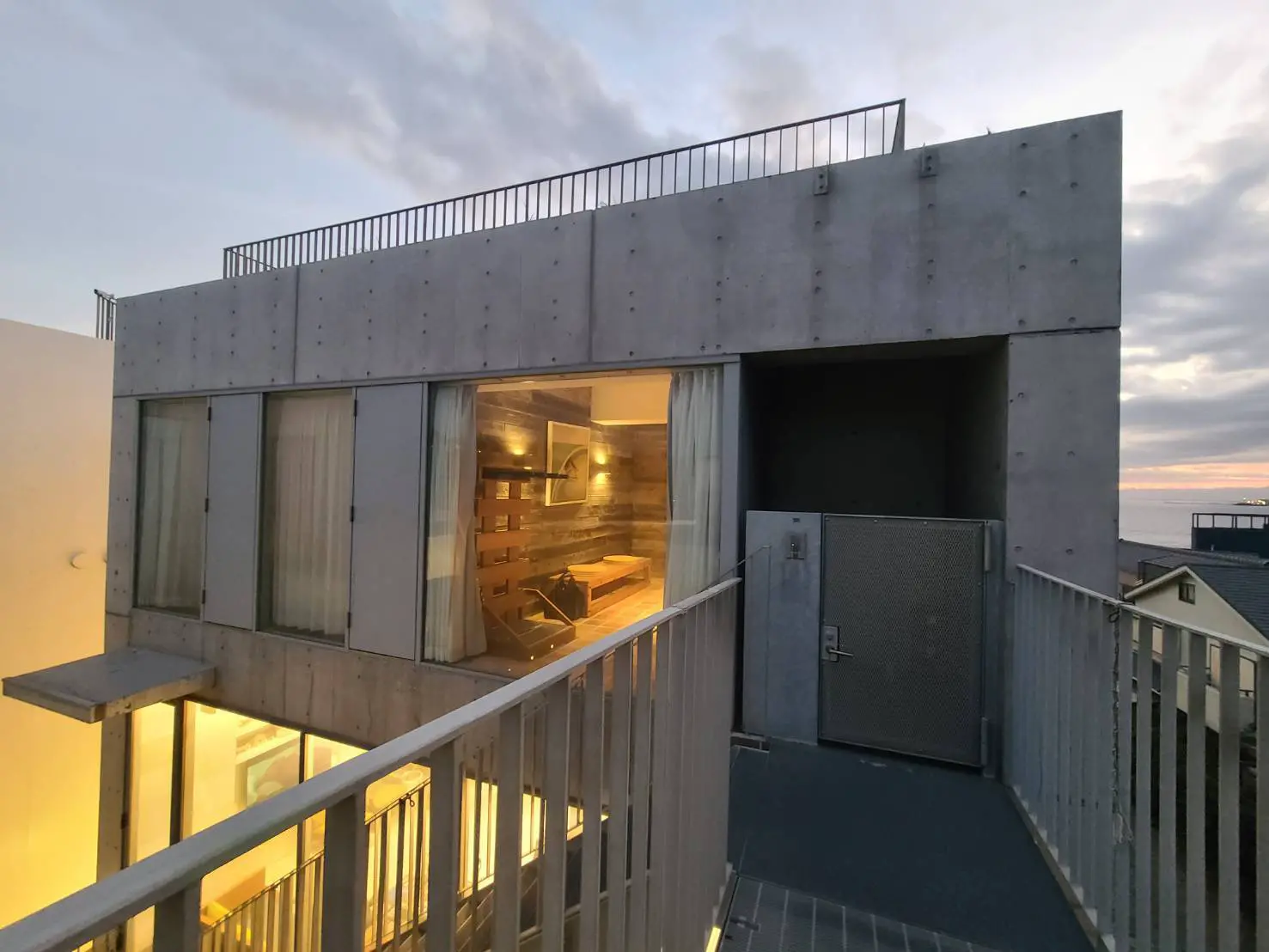 夕暮れ時に撮影されたコンクリート打ち放しの現代建築デザインの家、大きなガラス窓から室内照明が漏れる外観