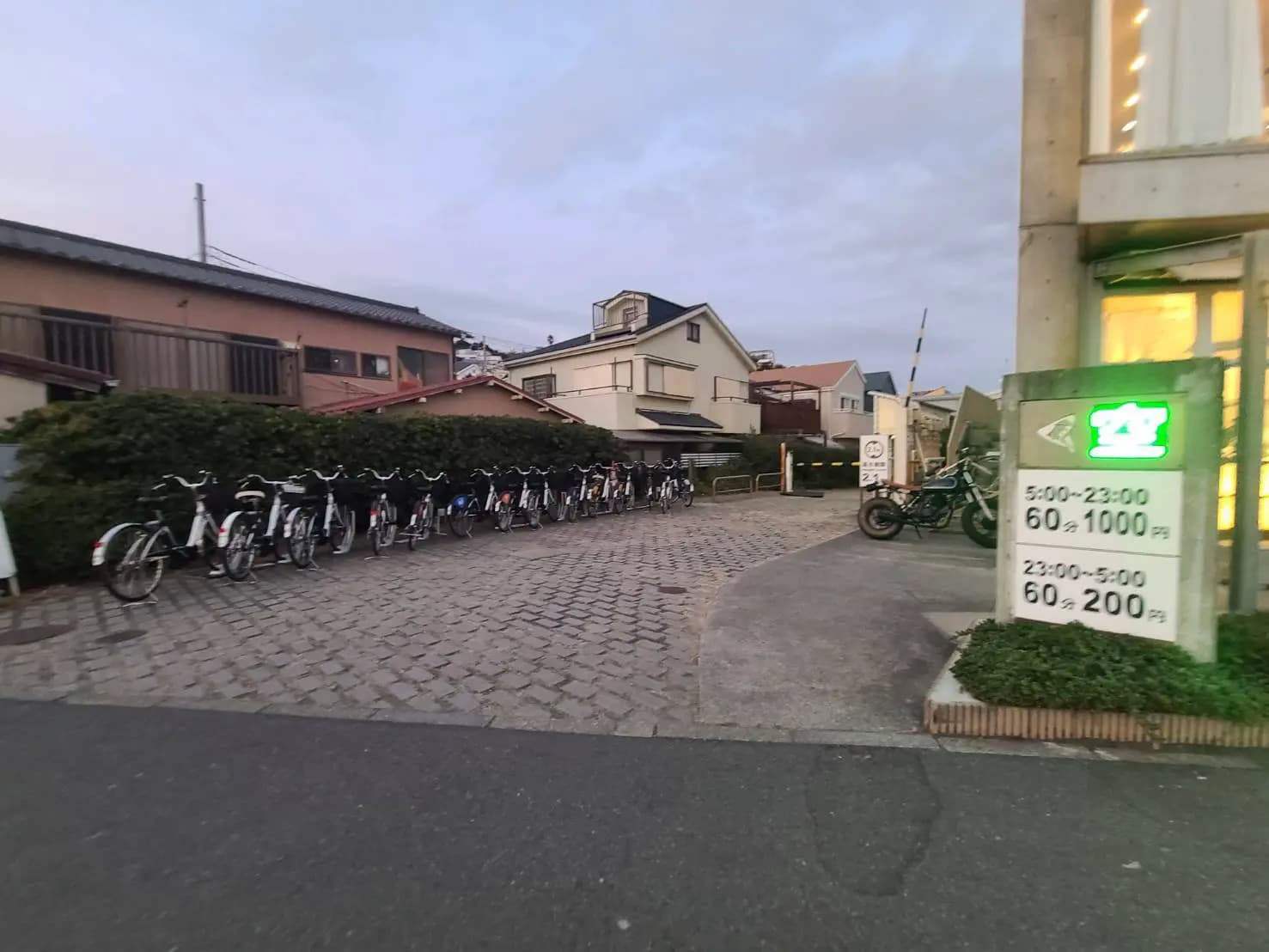 夕暮れ時の駐輪場、整然と並んだ自転車、コンクリートの舗道、背景に住宅街、24時間営業の標識が目立つ街角