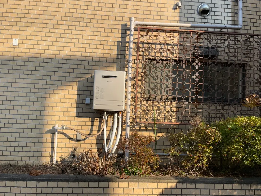 壁に取り付けられたガス給湯器と配管、住宅の外観、格子状の保護カバーが付いた窓