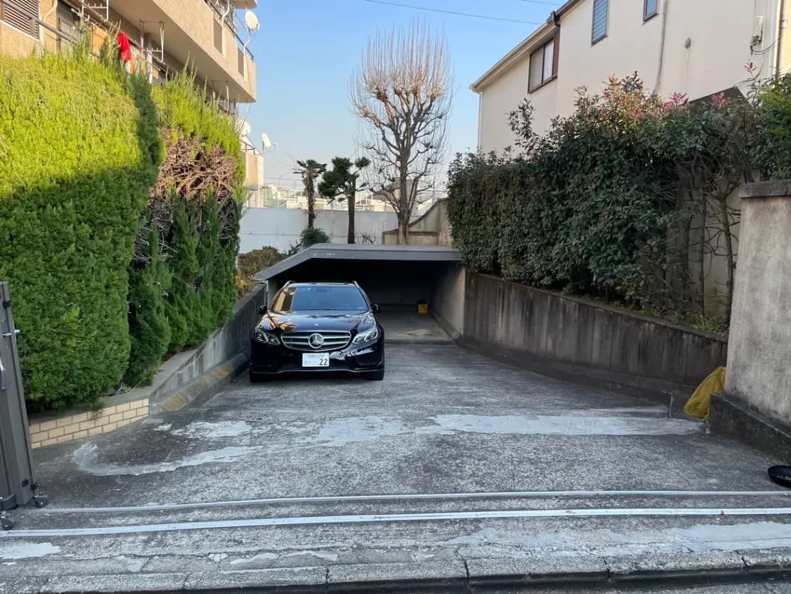 都市の一戸建て住宅のカーポートに駐車されている黒い高級車、周囲には緑豊かな樹木と明るい日差し
