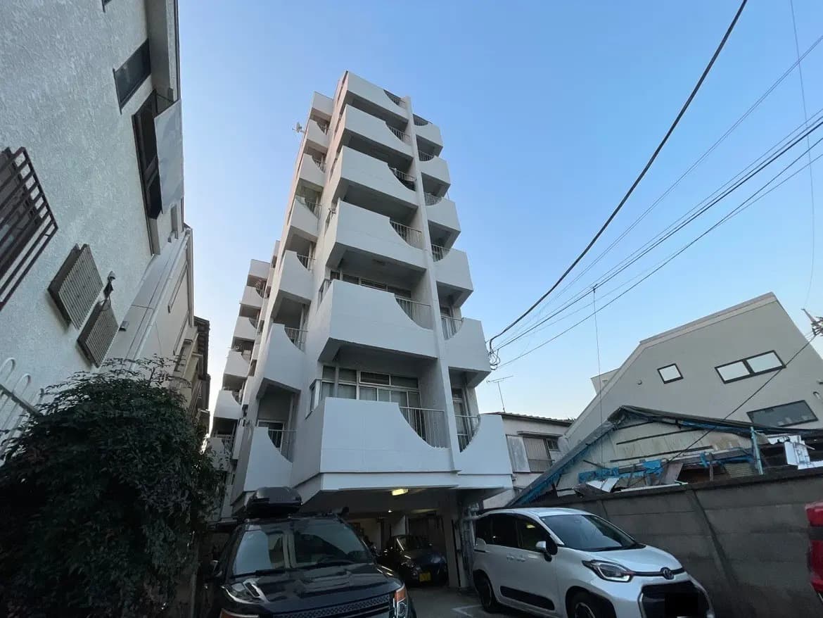 都市の住宅街にある白い多階建てアパート、青空と電線、駐車場に停車中の車