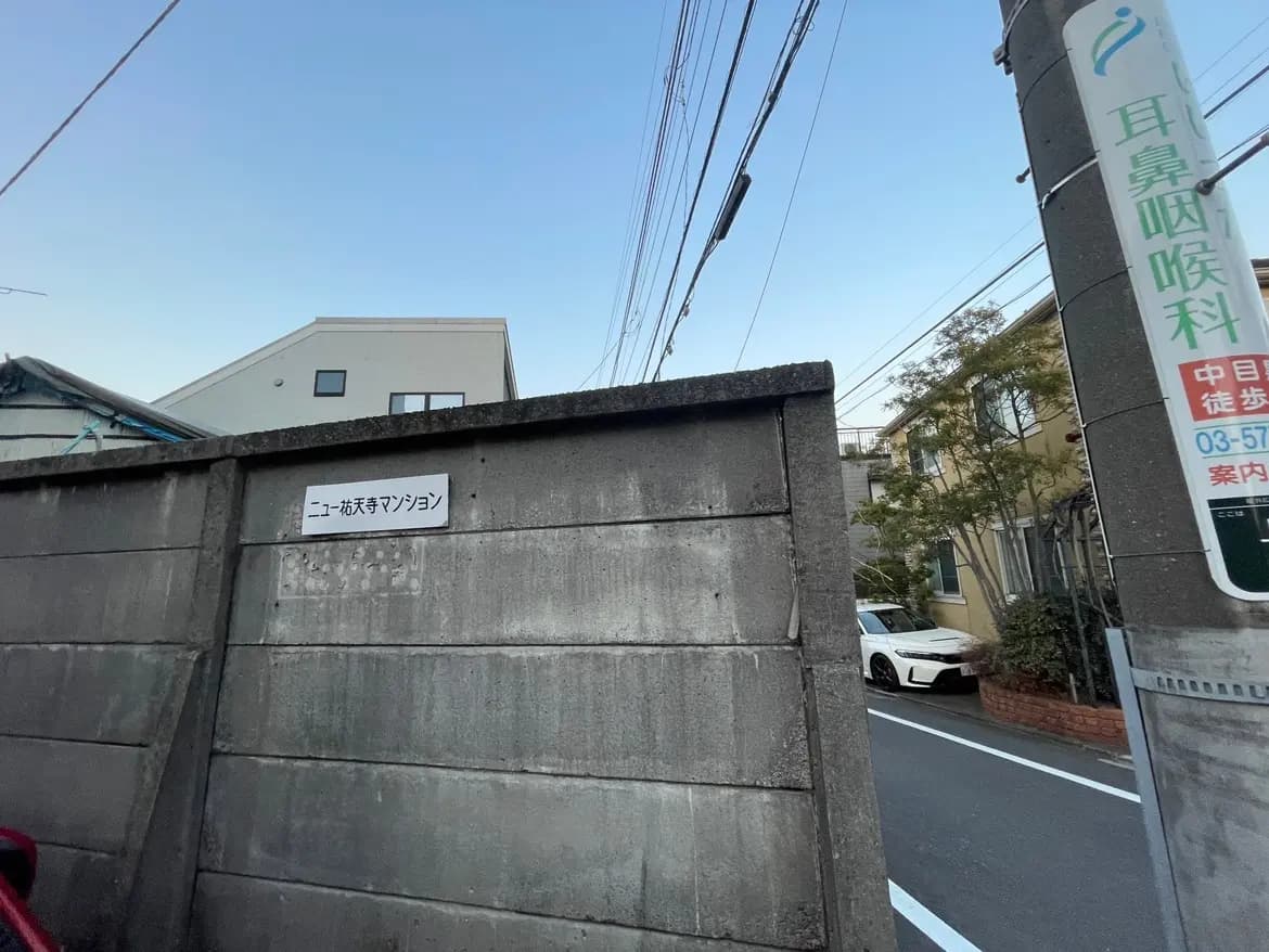 コンクリート製の塀に貼られた「ニートが家デマンション」の看板が見える街角、背景に住宅と車、道路標識がある風景