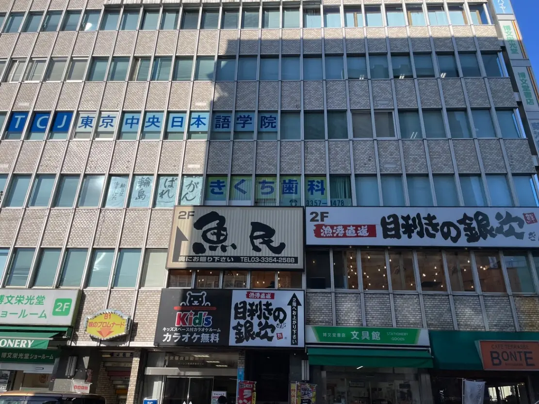 日中に撮影された都市の建物の外観、様々な店舗の看板が並び、青空が見える