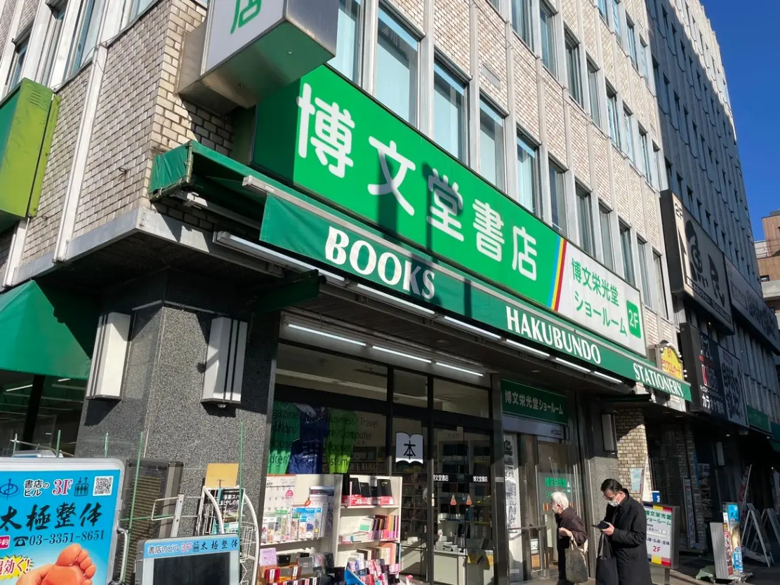 日差しが照りつける青空の下、街の本屋「HAKUBUNDO」の店舗外観、歩道に人々