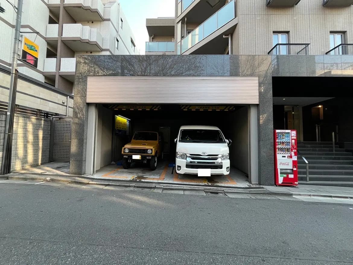 都市のマンションの駐車場入り口、自動車二台と自動販売機が見える外観