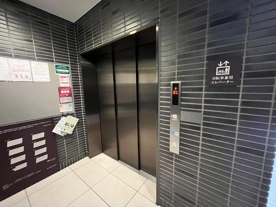 建物内エレベーター前、階表示灯がB1を照らす、壁に掲示物と案内標識がある現代的なデザイン