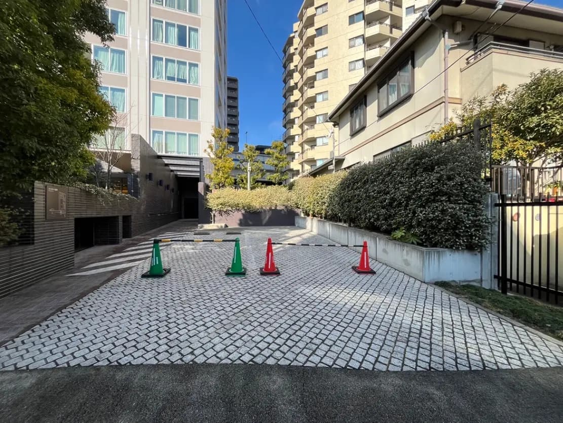 都市のマンションとアパートに挟まれた舗装された道路、緑の植栽と赤緑の交通コーンが配置されたエントランス