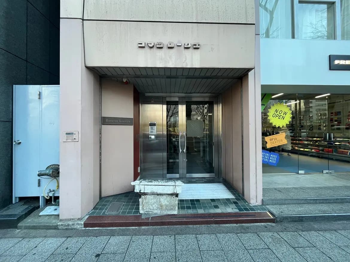 ビジネスビルの入り口、ガラス扉とサインボード、「NOW OPEN」の宣伝看板が見える都市の風景