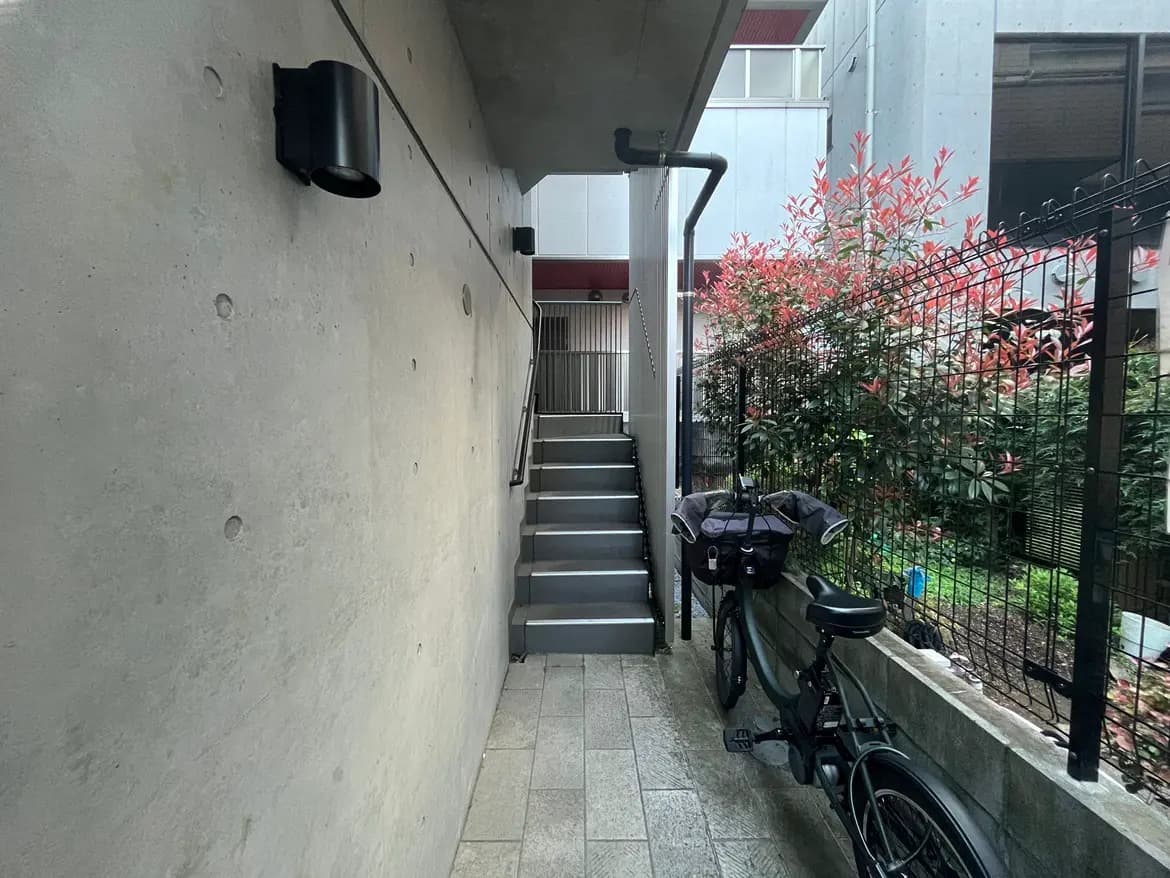 - コンクリートの壁と階段、横に置かれた自転車、周囲の植物が豊かなエントランスの風景